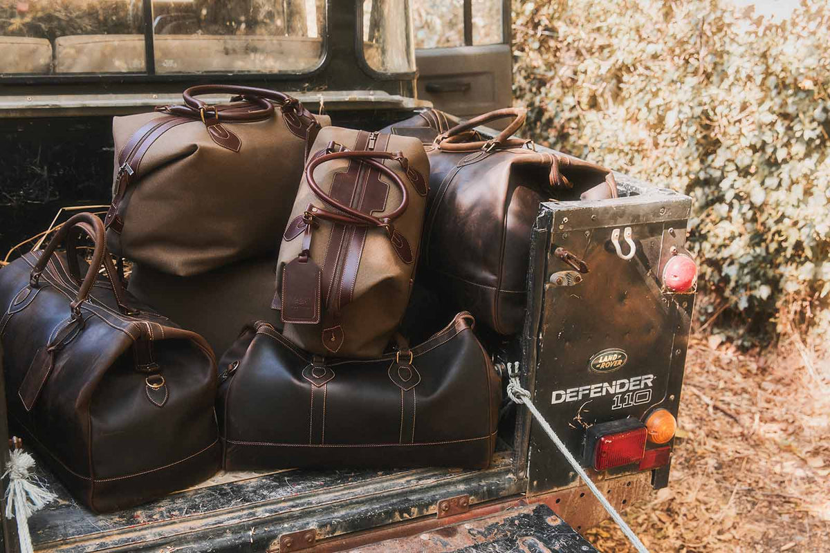 The Tusting Como Collection of Explorer Luggage, set next to a beautify, dark blue Aston Martin car